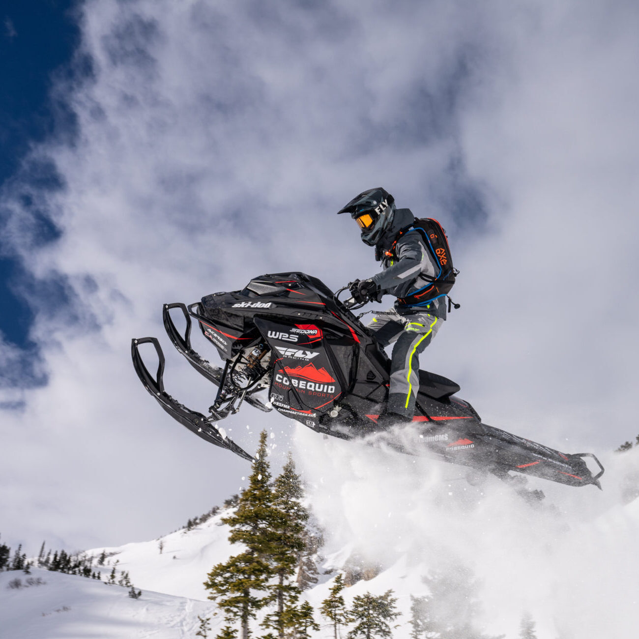 A man on a black snowmobile soars through the air against a cloudy blue sky, captured mid-jump with snow-covered mountains and pine trees in the background. He wears a black helmet and winter gear, leaning forward as the snowmobile tilts slightly upward.