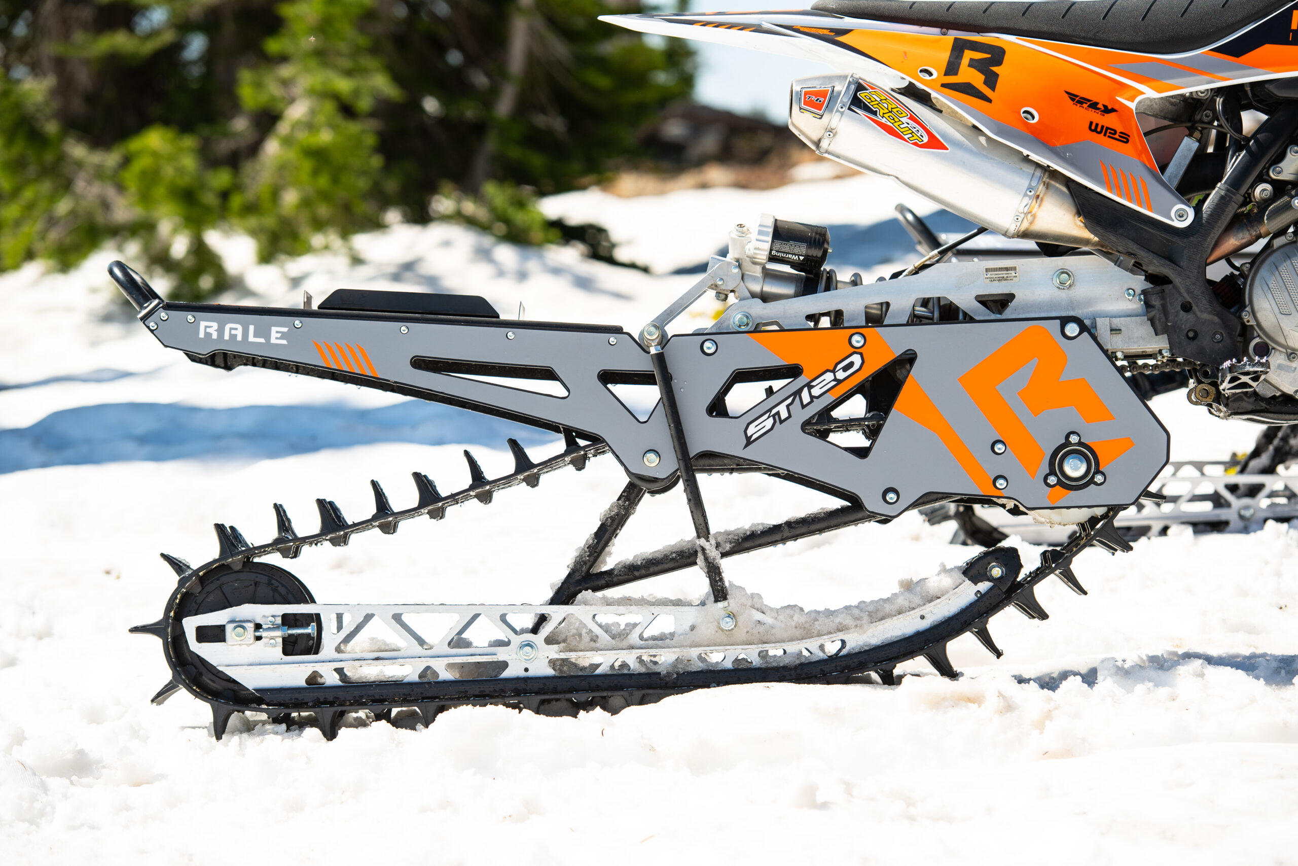 Rale Industries-009 A man on a black snowmobile soars through the air against a cloudy blue sky, captured mid-jump with snow-covered mountains and pine trees in the background. He wears a black helmet and winter gear, leaning forward as the snowmobile tilts slightly upward.